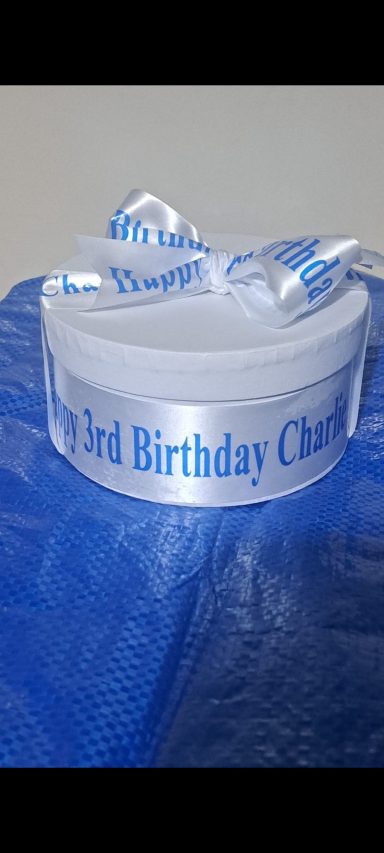 White birthday cake box with blue "Happy 1st Birthday" ribbon, resting on a blue surface.